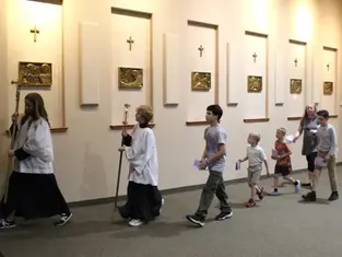 Children and adults walk in procession along a church wall with gold plaques and crosses. They carry papers, creating a solemn atmosphere.