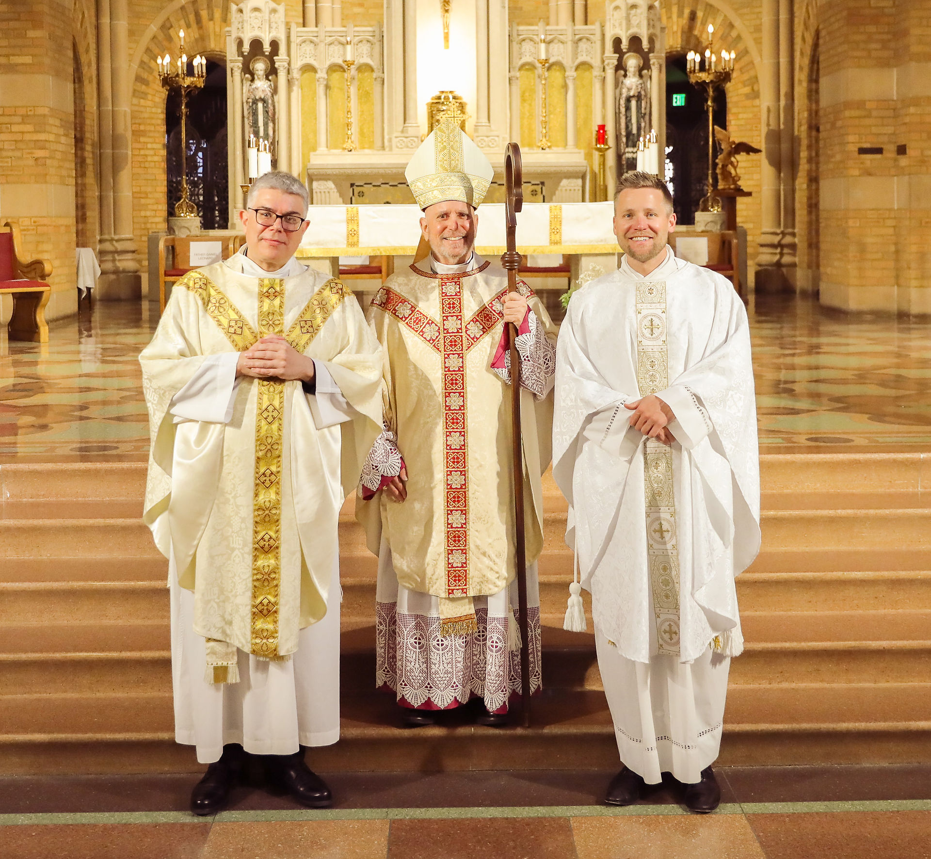 Two priests stand for a photo with an archbishop in an ornate chapel following a religious ceremony.