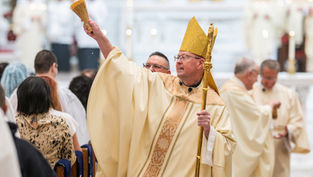Bishop in gold vestments and mitre raises an aspergillum during a church ceremony, with congregation and clergy blurred behind.