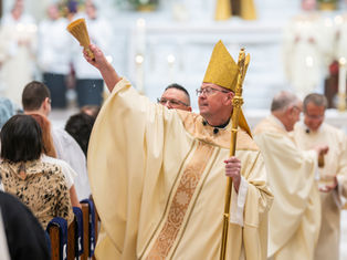 Bishop in gold vestments and mitre raises an aspergillum during a church ceremony, with congregation and clergy blurred behind.