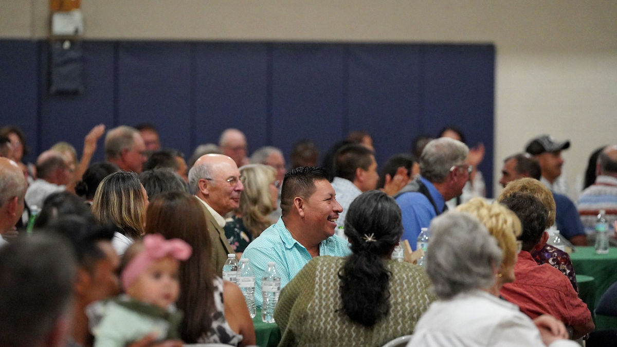 People seated at tables in a crowded hall, having conversations. The backdrop is blue, and the mood is lively. Water bottles on tables.