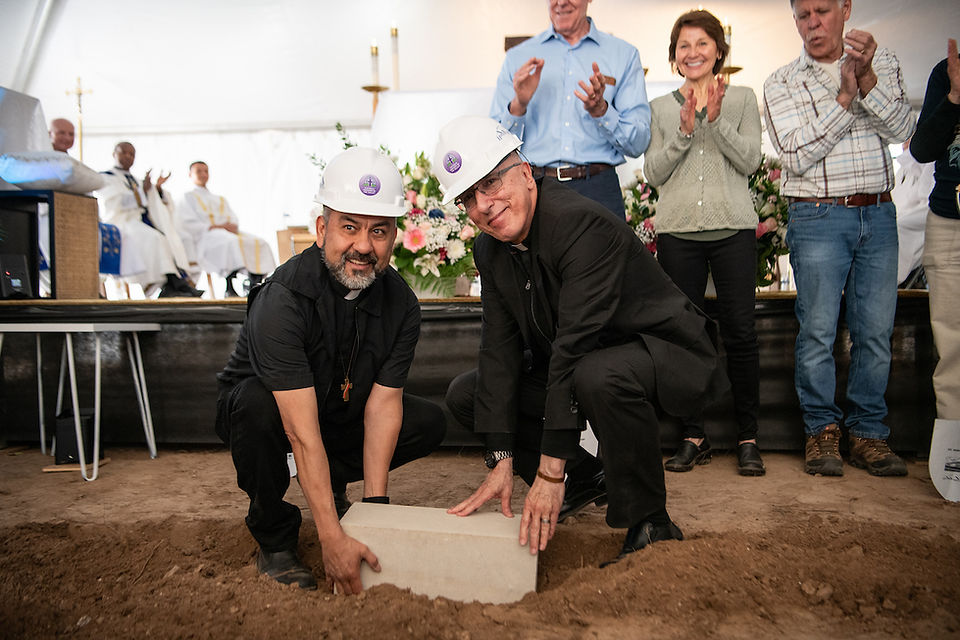 Two clergy members wearing hard hats place a concrete cornerstone block into the ground during the ceremony.