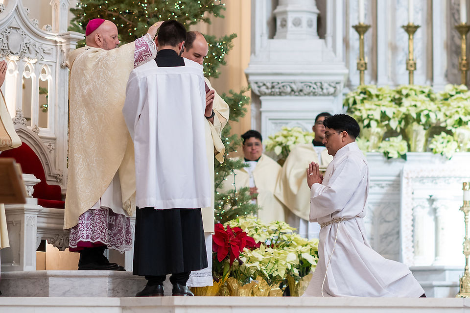 A bishop in ornate robes blesses a kneeling man in white during a church ceremony, with priests and festive poinsettias in the background.