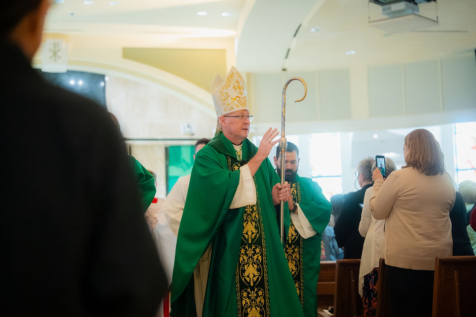 Bishop in green vestments and mitre holds a crozier during a church service while a woman films on her phone.