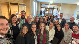 Smiling group selfie of coworkers in a brick hallway near an EXIT sign, posing closely in a bright indoor lobby