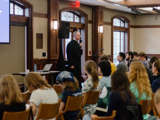 A speaker addresses an attentive audience in a wood-paneled room. A laptop and projector screen are visible, creating an academic atmosphere.