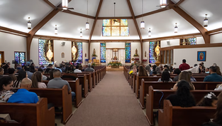 People seated in a church with stained glass windows, wooden pews, and lit chandeliers. A peaceful and solemn atmosphere.