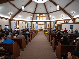 People seated in a church with stained glass windows, wooden pews, and lit chandeliers. A peaceful and solemn atmosphere.