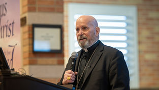 Bald man in a black suit speaks into a microphone at a podium. Text on the wall includes "Jesus Christ". Brick background.