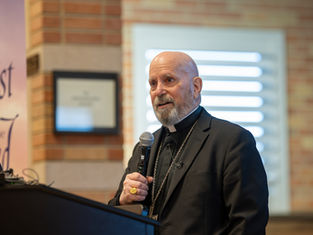 Bald man in a black suit speaks into a microphone at a podium. Text on the wall includes "Jesus Christ". Brick background.