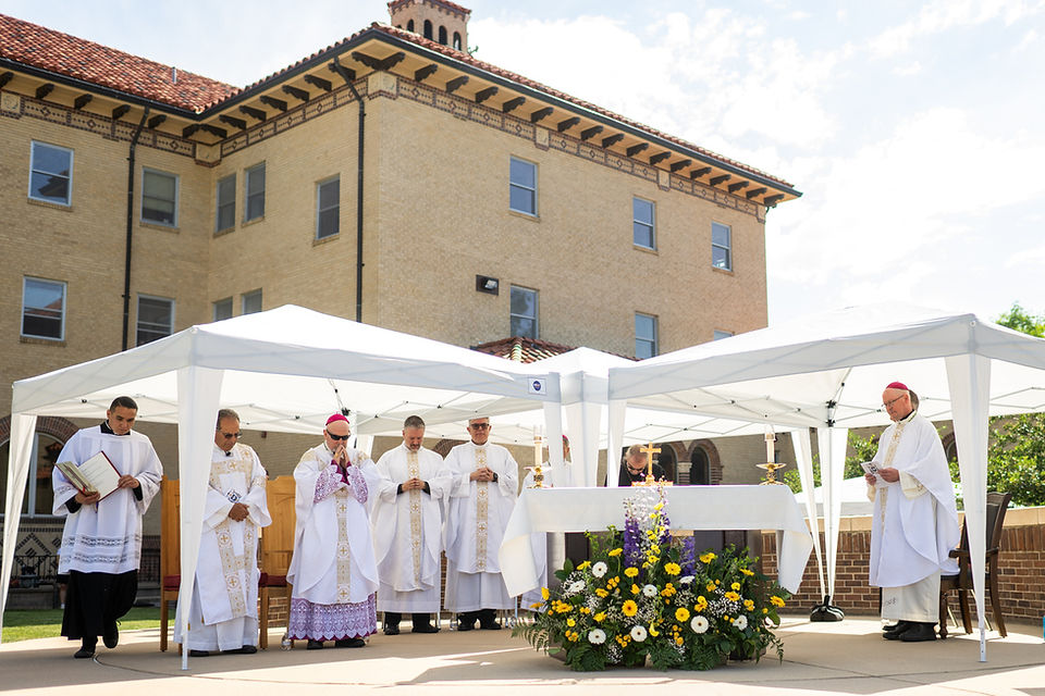 Outdoor Catholic Mass with priests in white vestments under canopies beside a flower-decorated altar and brick building.