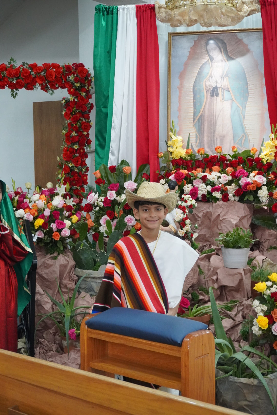 Boy in straw hat and striped shawl smiles in front of vibrant flowers and Mexican flag colors, with a Virgin of Guadalupe image above.