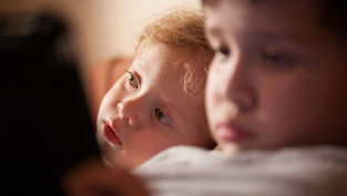 Two young children in warm indoor light, one blond child gazing upward while the other sits blurred in the foreground.