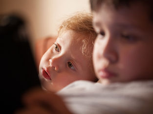 Two young children in warm indoor light, one blond child gazing upward while the other sits blurred in the foreground.