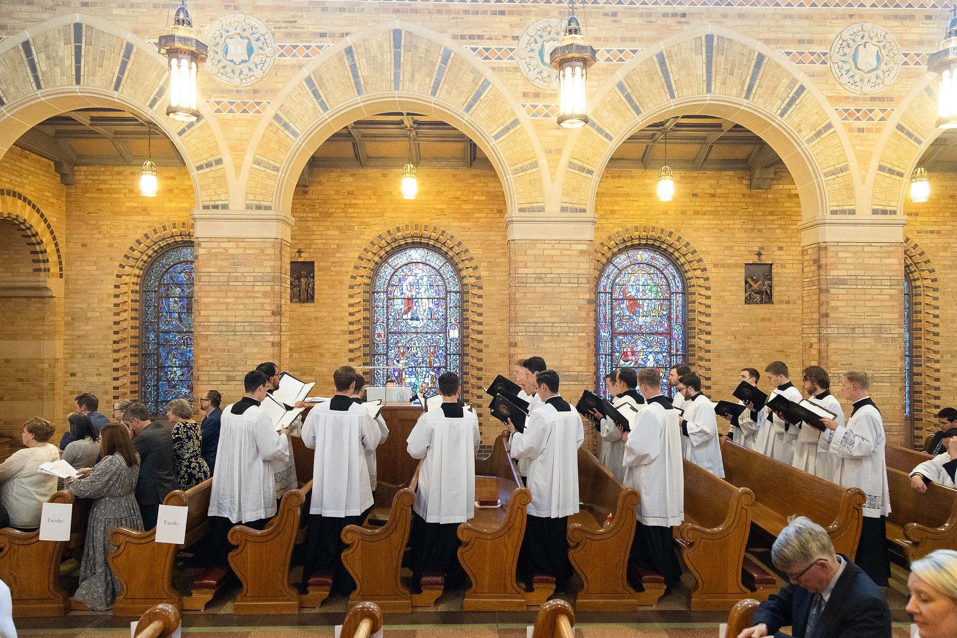 Seminarians in robes stand in the seminary chapel, leading the congregation in song during a religious service. The background shows the ornate stained glass windows and beautiful brickwork of the chapel.