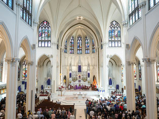 Crowded cathedral service with congregation standing in a bright Gothic church, stained glass, white arches, and purple altar drapes