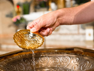 Hand pours water from an ornate gold shell cup into a basin in a softly lit interior, with warm reflective tones.