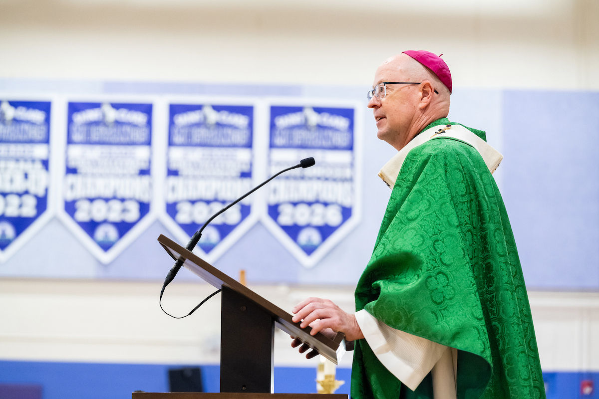 Bishop in green vestments speaks at a podium in a church gym, with blue banners reading 2022 and 2026 behind him.