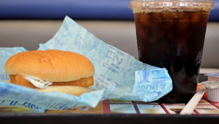 Fish sandwich on blue wrapper, iced cola in plastic cup with lid and straw on tray. Restaurant setting, casual dining vibe.