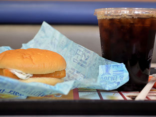 Fish sandwich on blue wrapper, iced cola in plastic cup with lid and straw on tray. Restaurant setting, casual dining vibe.