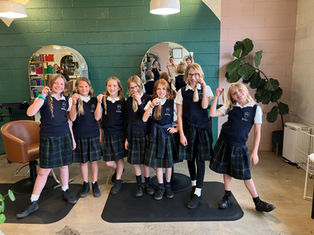Seven girls in navy uniforms hold cut hair in a salon with green and pink walls. They smile, showing pride and excitement.