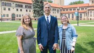 Three smiling adults stand on a grassy campus lawn before a historic brick building with red roofs and a cross, sunny and formal.