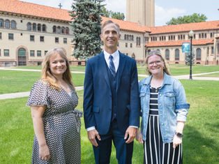 Three smiling adults stand on a grassy campus lawn before a historic brick building with red roofs and a cross, sunny and formal.