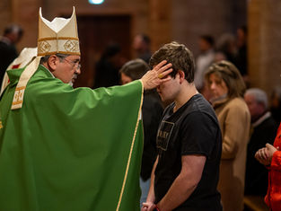 A bishop in green robes blesses a young man in a church, touching his forehead. The congregation is visible in the background.
