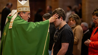 A bishop in green robes blesses a young man in a church, touching his forehead. The congregation is visible in the background.