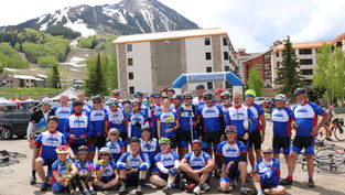 Cycling team in blue jerseys poses under a The Denver Post Ridetherockies arch, with snowy mountain and lodges behind them.