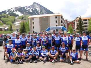 Cycling team in blue jerseys poses under a The Denver Post Ridetherockies arch, with snowy mountain and lodges behind them.