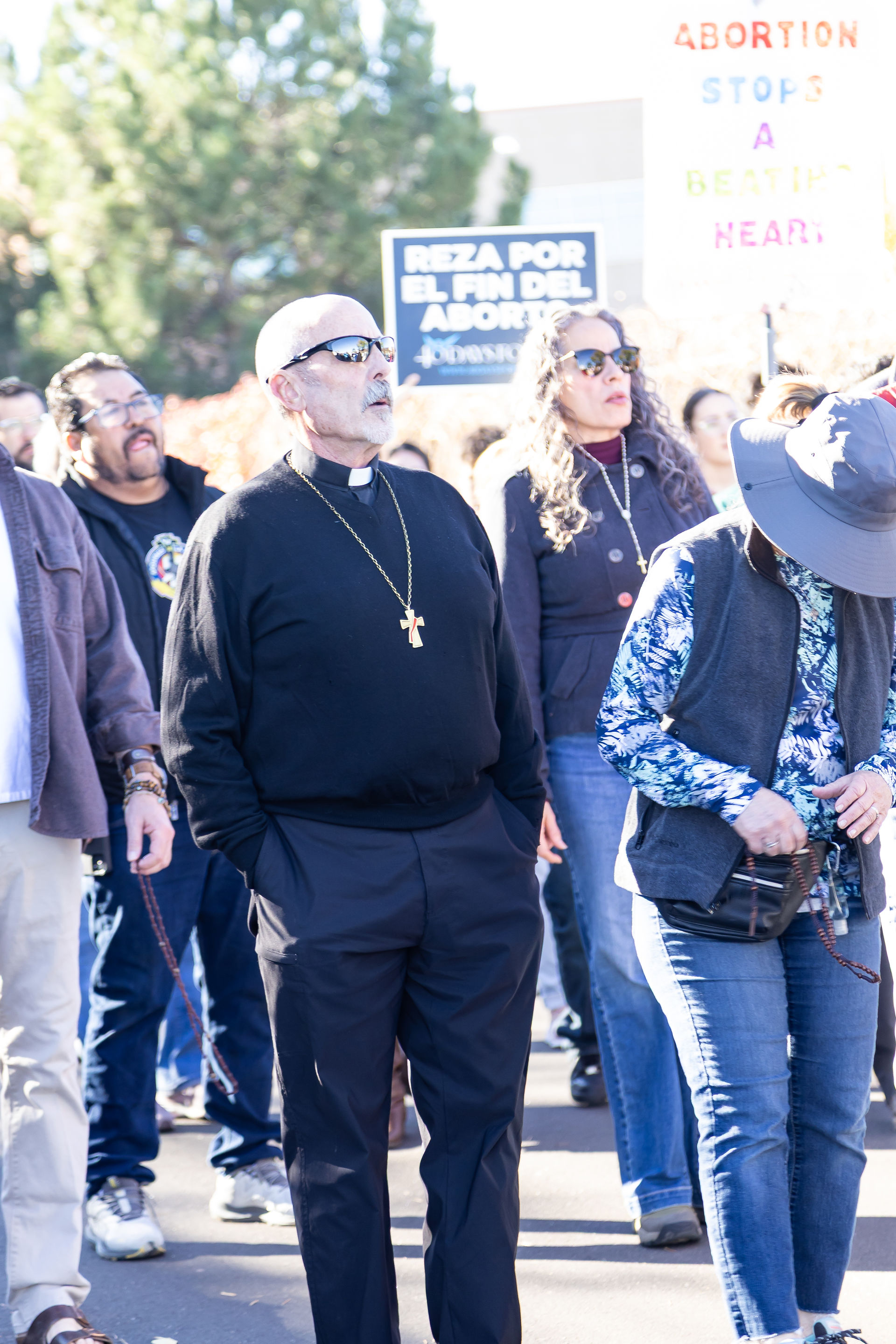 People in a procession hold signs advocating for life. A deacon dressed in black leads. Background shows trees and bright sunlight.