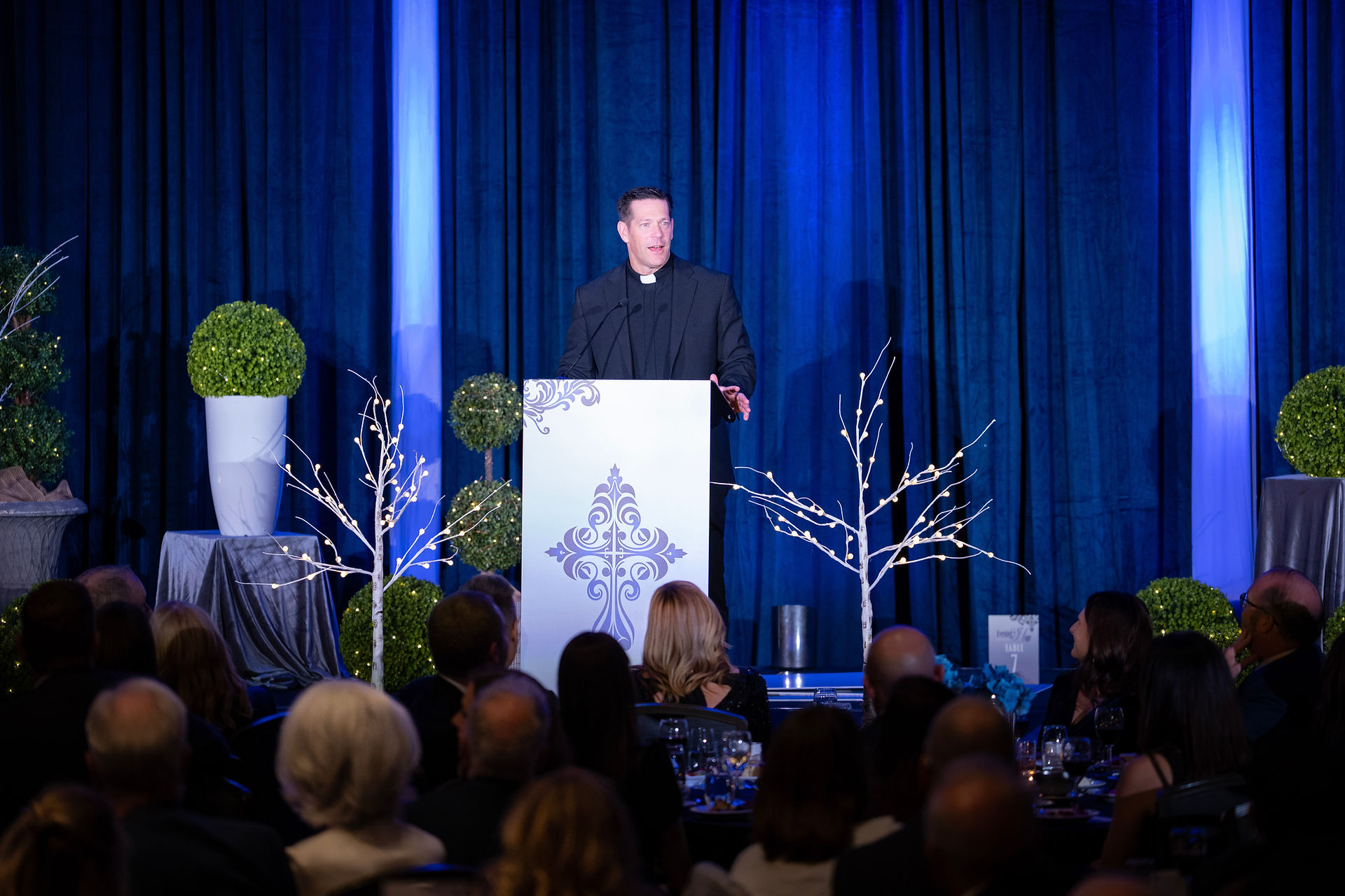 A man speaks at a podium with decorative patterns, set against blue curtains. Audience listens, surrounded by lit branches and green plants.
