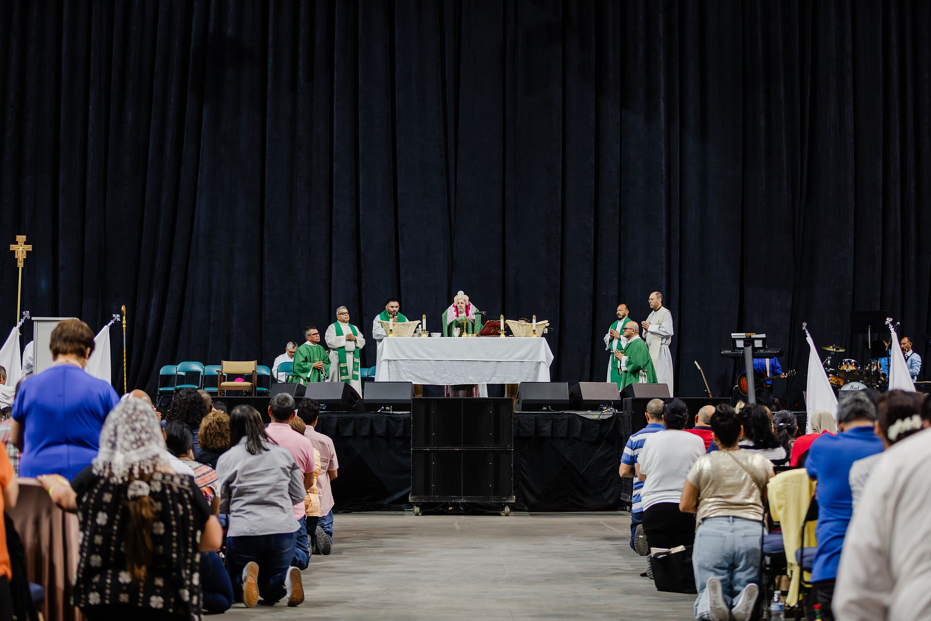 Clergy in green robes lead a church service on stage. Attendees kneel in prayer in a large hall, with dark curtains in the background.