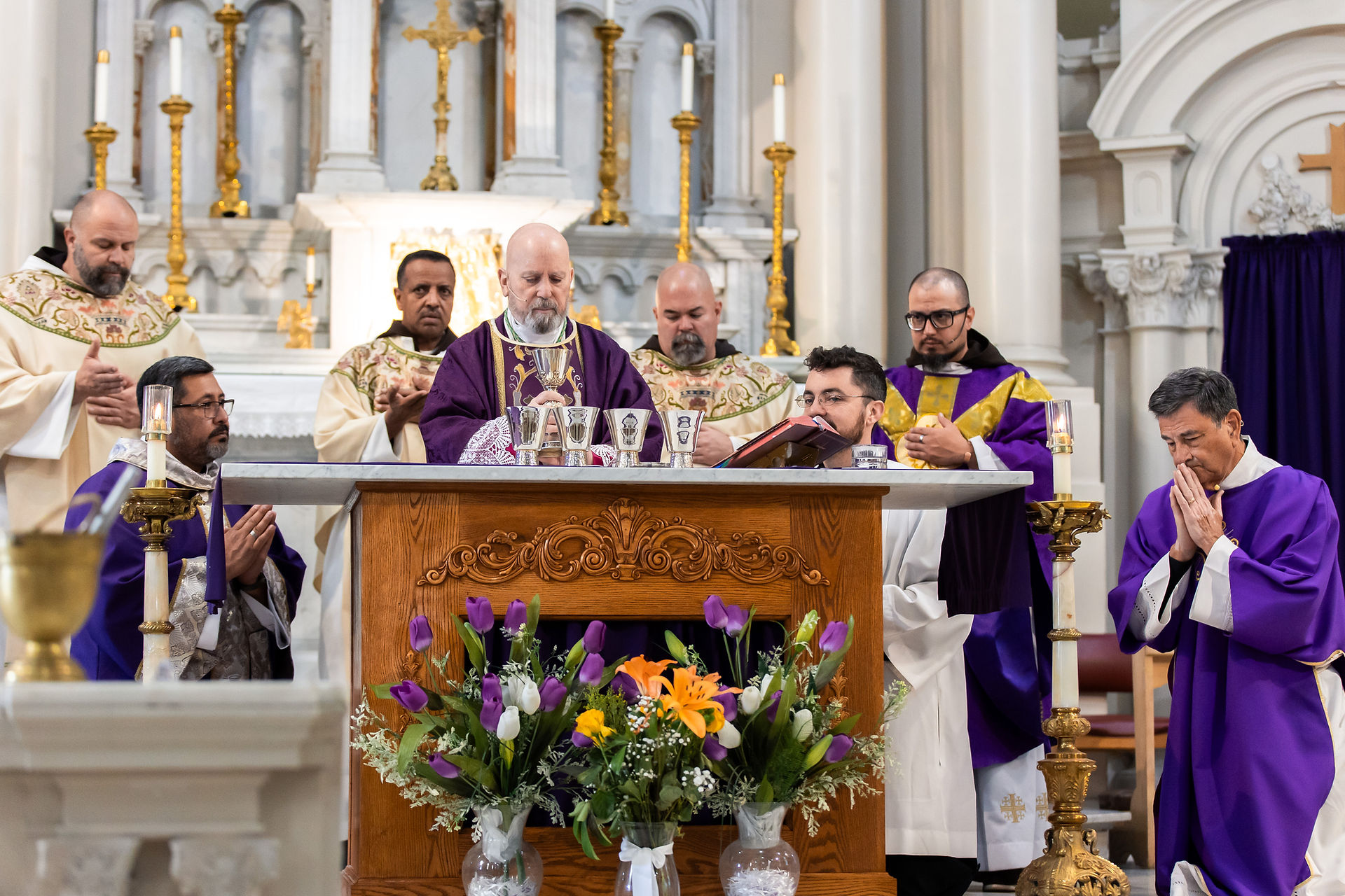 Clergy in purple robes perform a religious ceremony at an ornate altar with flowers and candles. One priest holds a chalice.
