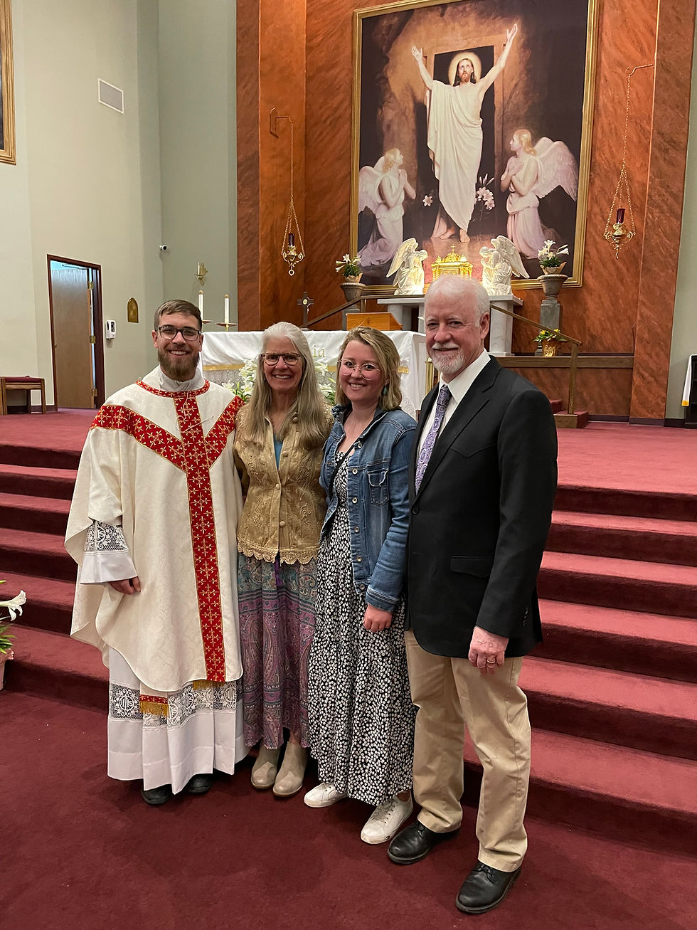 Four people pose in a church before a Jesus artwork. One wears religious attire, others in casual clothes. Red carpet and altar in background.