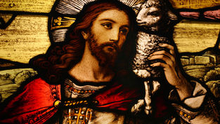 Stained glass depicts a bearded man with a halo holding a lamb, wearing red and white robes. The scene is serene with a rural backdrop.