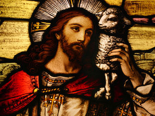 Stained glass depicts a bearded man with a halo holding a lamb, wearing red and white robes. The scene is serene with a rural backdrop.