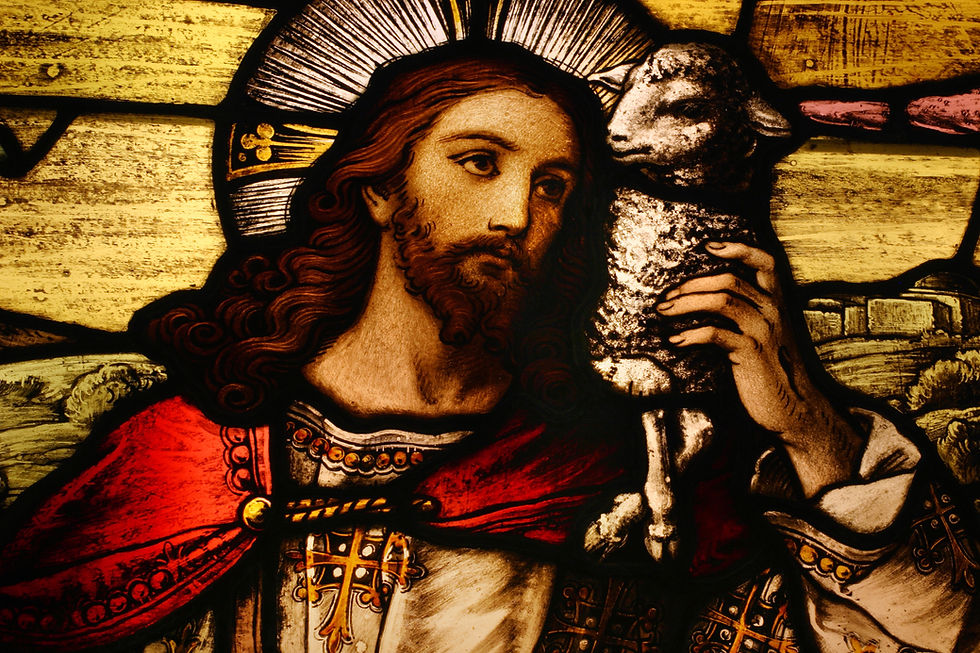 Stained glass depicts a bearded man with a halo holding a lamb, wearing red and white robes. The scene is serene with a rural backdrop.