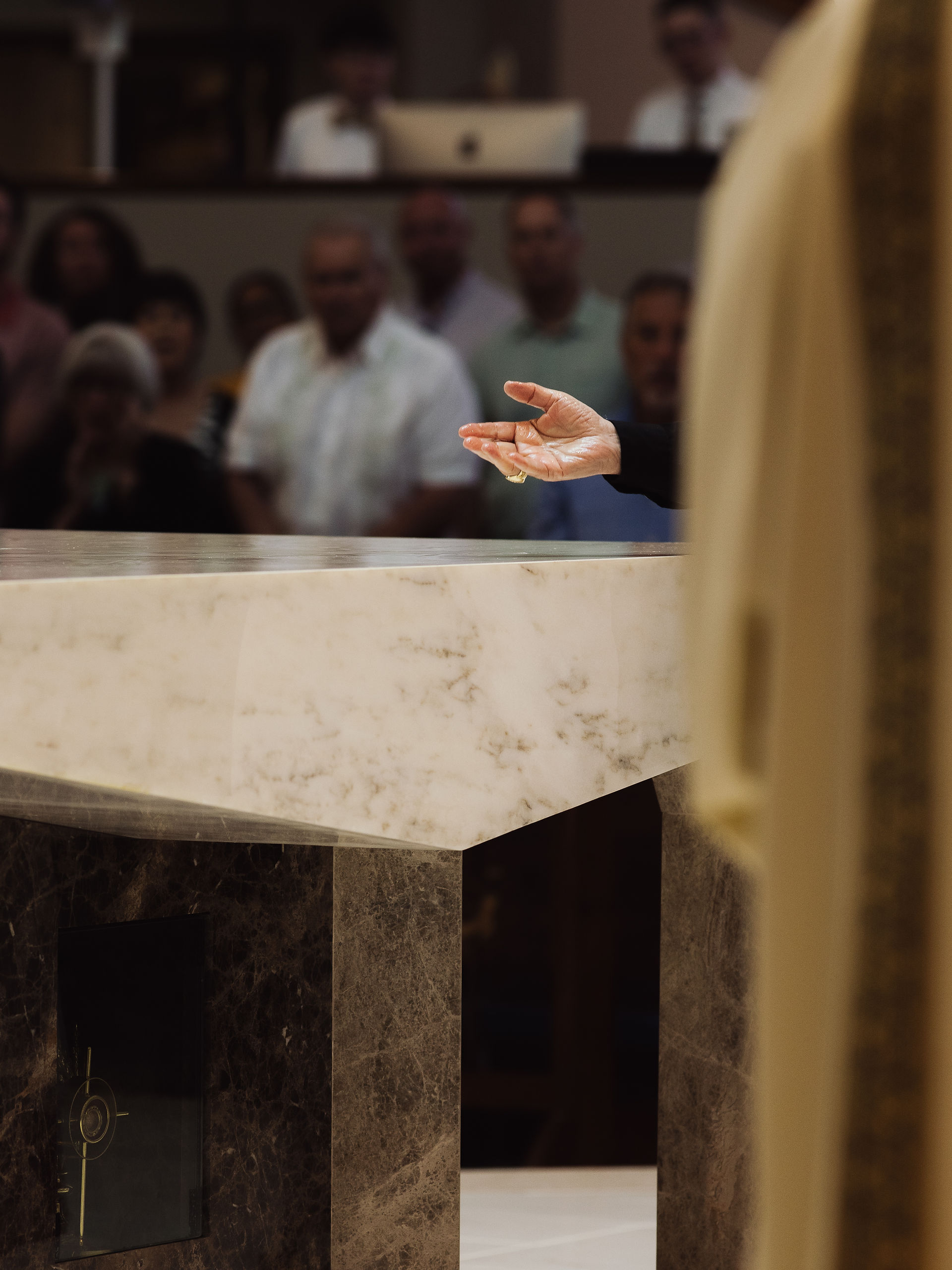 A hand gestures over a marble altar in a church. People are in the background. The scene is solemn, with muted colors and dim lighting.