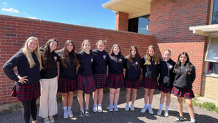 Ten girls in plaid skirts and black hoodies pose smiling outside a school building with brick walls under a clear blue sky.