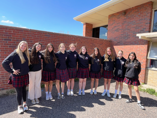 Ten girls in plaid skirts and black hoodies pose smiling outside a school building with brick walls under a clear blue sky.