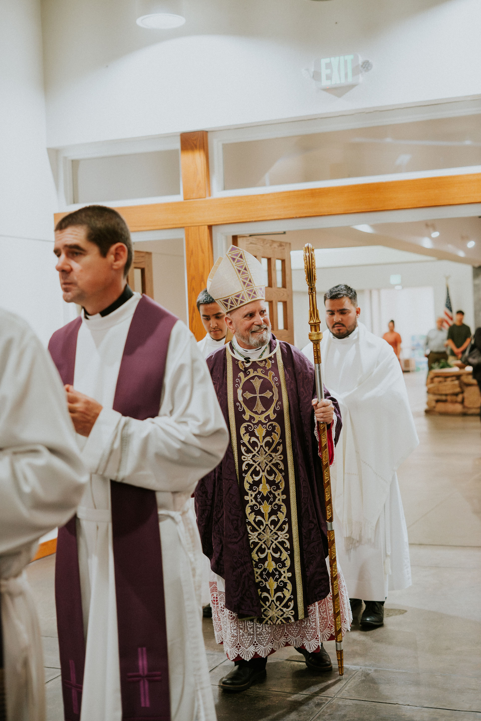 Clergy in ornate robes process through a church hallway. A bishop in purple holds a crosier, surrounded by other clergy in white. Exit sign visible.