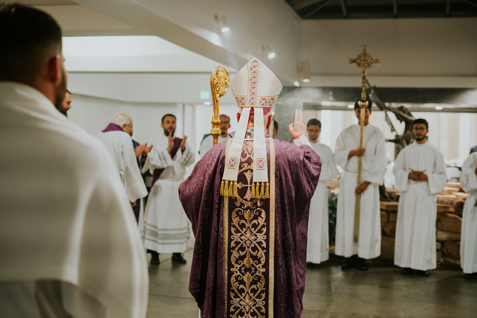 A bishop in ornate purple robes performs a blessing ceremony. Clergy in white robes stand in a circle inside a church, creating a solemn mood.