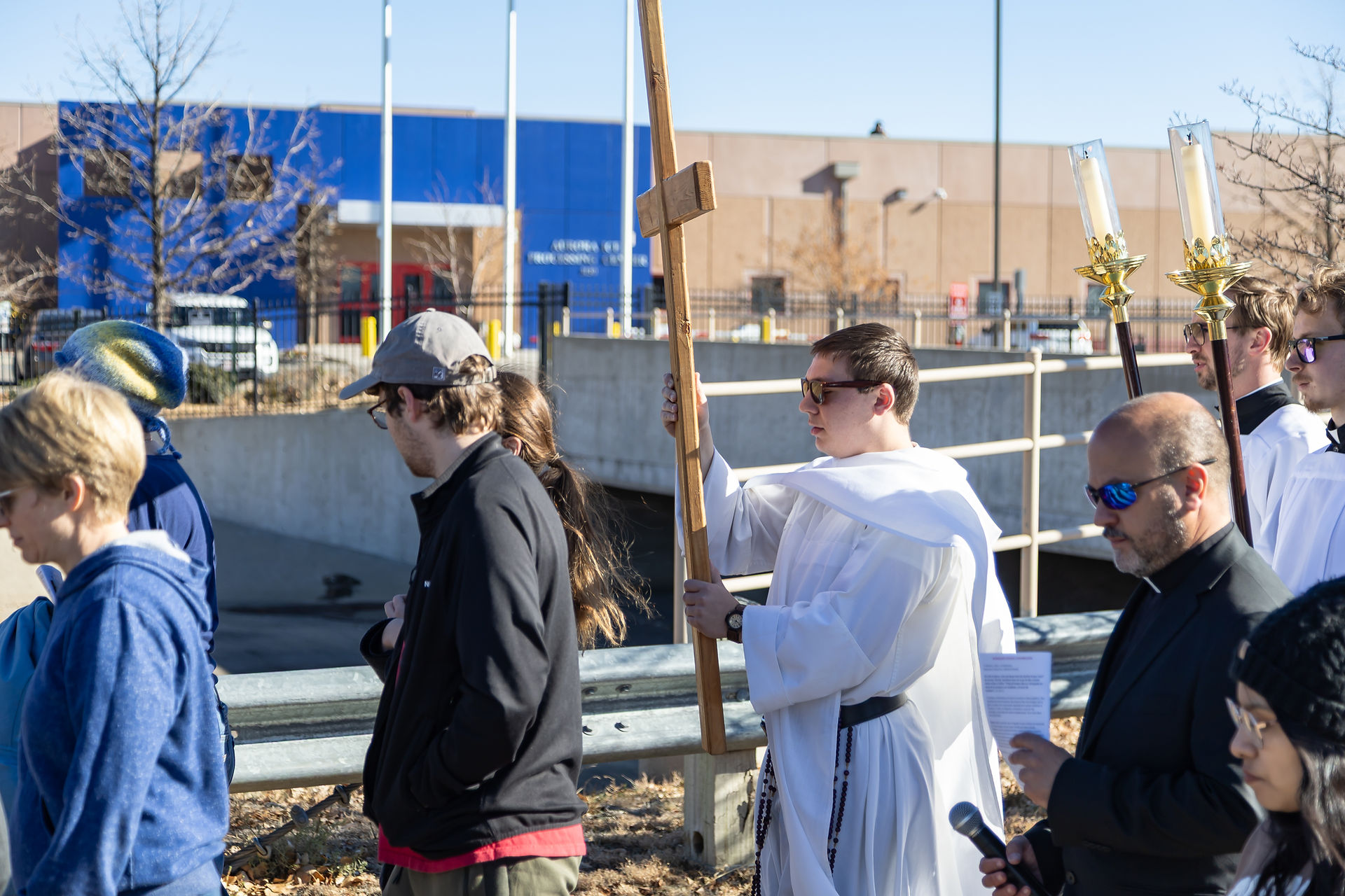 People in white robes carrying a cross and candles walk in a procession by a blue building. A man in black holds a microphone. Sunny day.