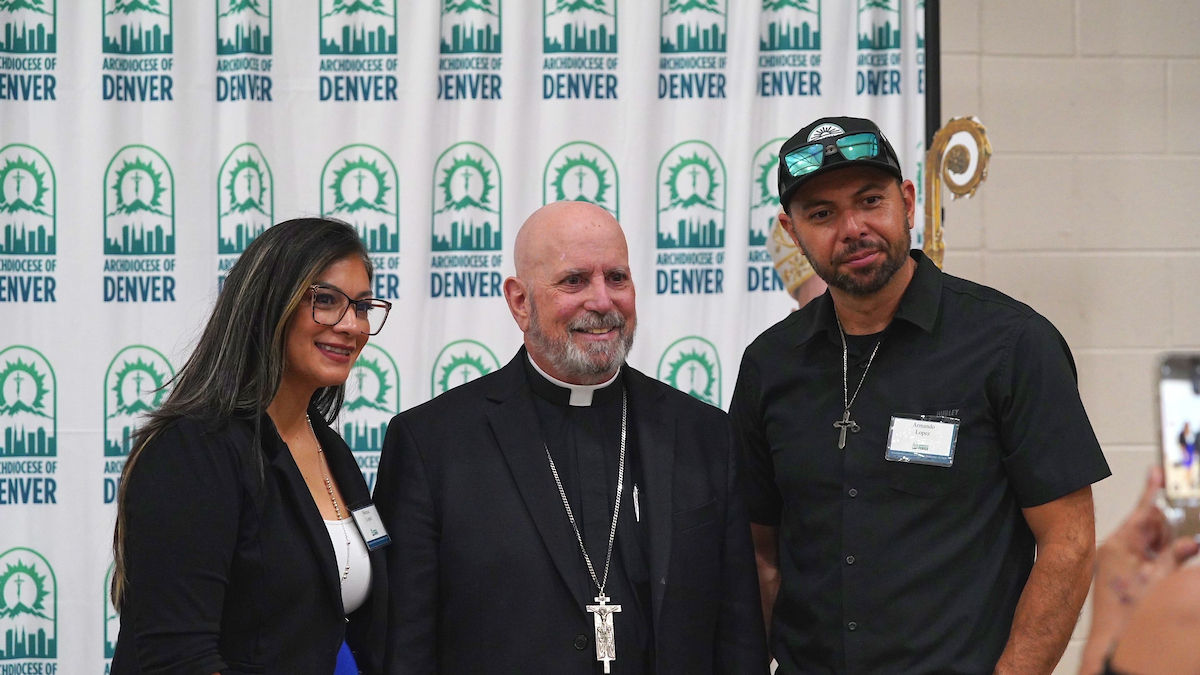 Three people pose smiling in front of an "Archdiocese of Denver" backdrop. The setting is bright and formal with a cheerful mood.