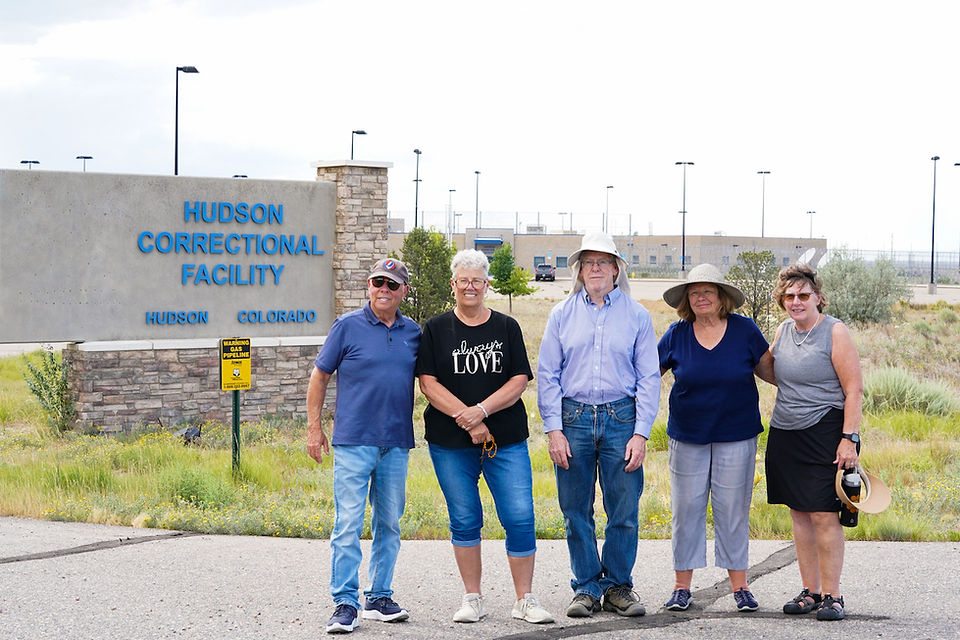 Five smiling adults stand outside Hudson Correctional Facility sign in Hudson, Colorado.