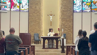 Priest at altar in church with a crucifix above, leading a service. Congregation stands in pews. Stained glass windows and brick wall backdrop.