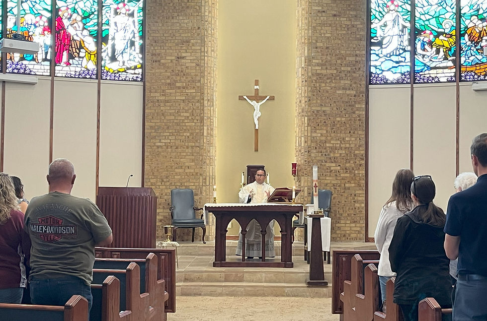 Priest at altar in church with a crucifix above, leading a service. Congregation stands in pews. Stained glass windows and brick wall backdrop.