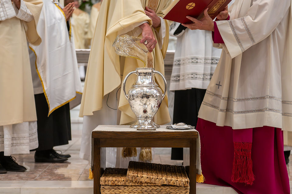 Clergy in cream robes pour liquid into a silver urn labeled SC. One holds a red book. Background shows more robes in a church setting.
